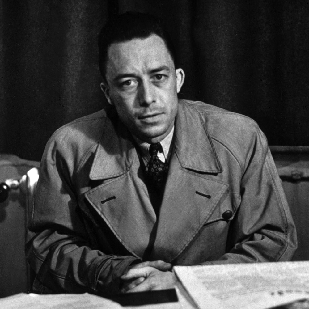 Photo en noir et blanc d’Albert Camus, écrivain et philosophe, avec une pile de ses œuvres majeures à ses côtés.