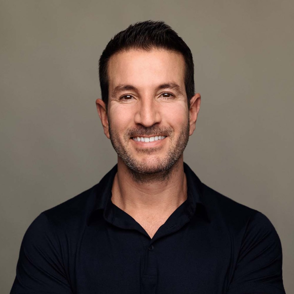 Photo d’Alex Michaelides, auteur de thrillers psychologiques, souriant à côté d’une pile de ses romans à succès.