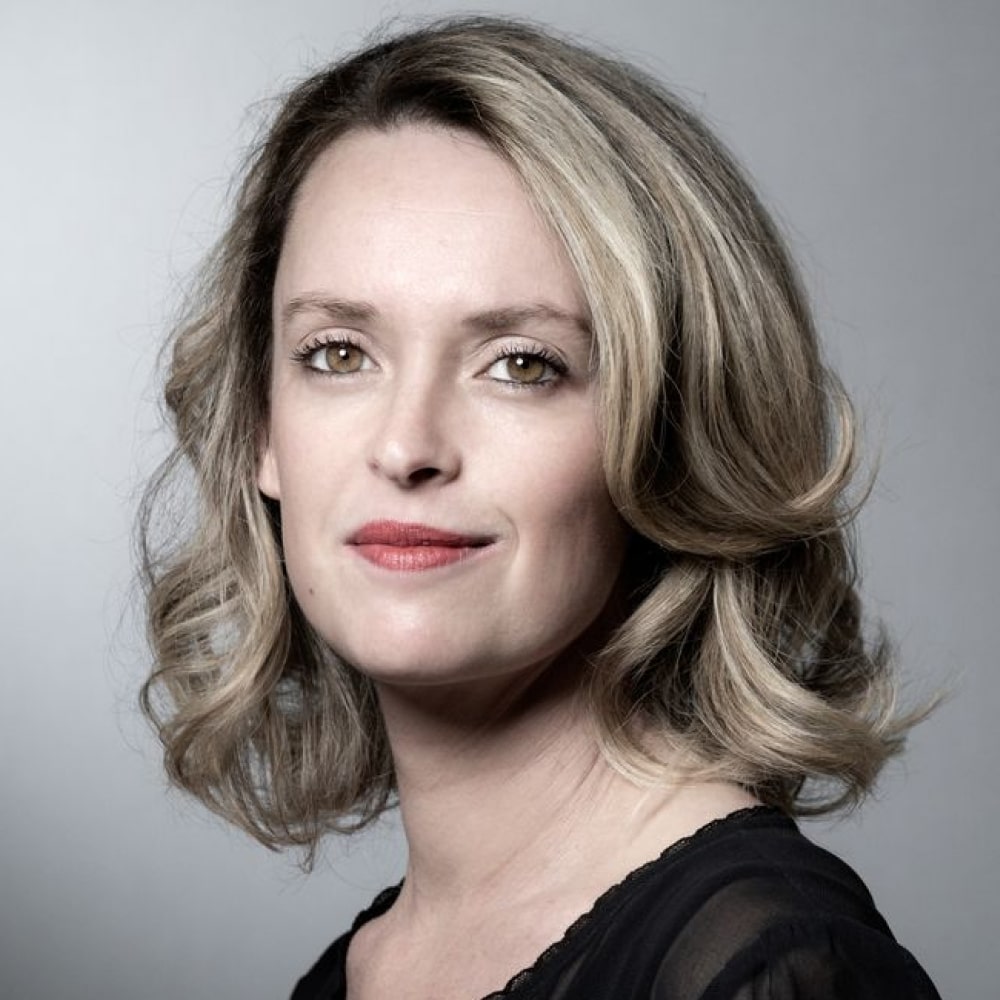 Portrait d’Aurélie Valognes, autrice de romans feel-good, posant devant une pile colorée de ses livres à succès.