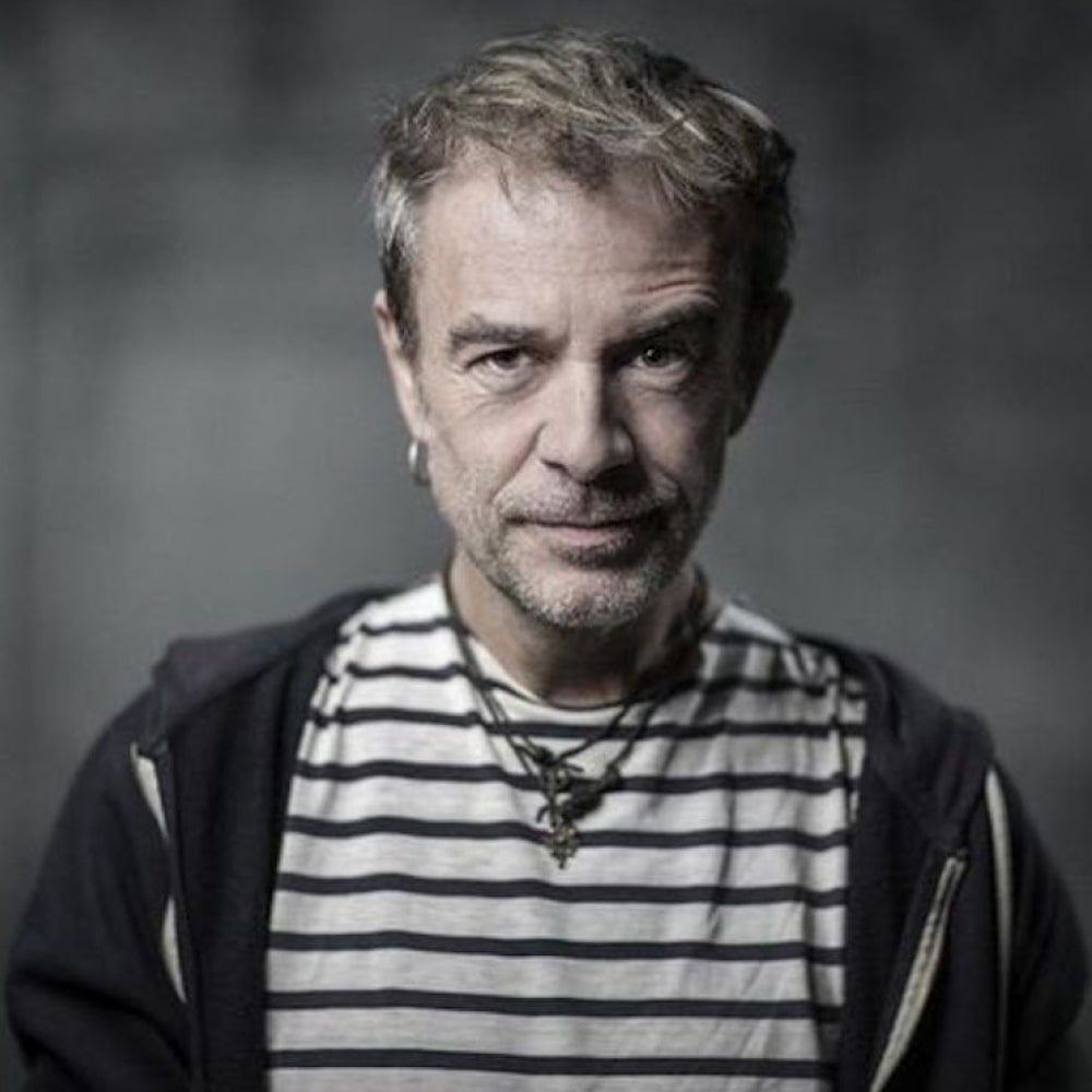 Portrait de Fabrice Caro, alias Fabcaro, romancier et dessinateur, souriant à côté d’une pile de ses romans pleins d’humour absurde.