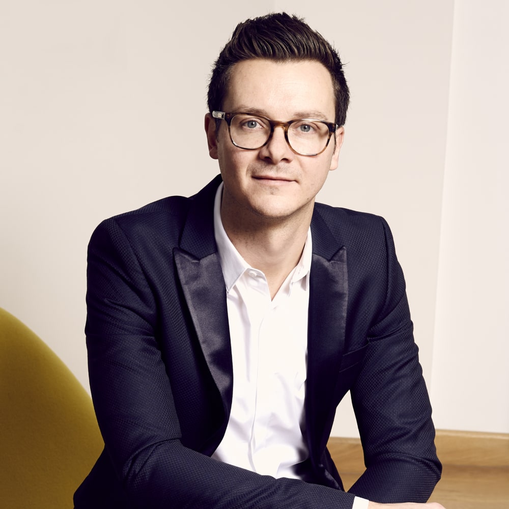 Photo de Julien Sandrel, romancier français, souriant à côté d’une pile de ses romans feel-good pleins d’émotion.