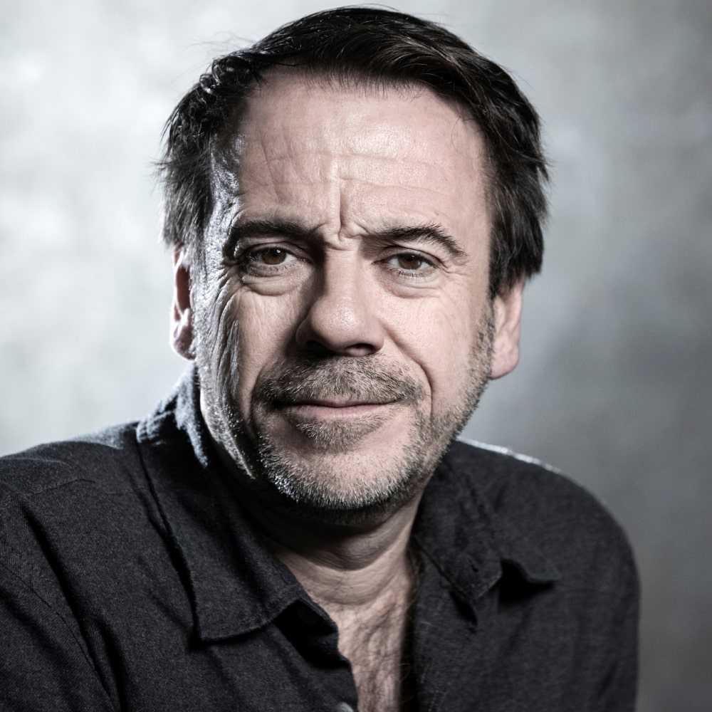 Photo de Michel Bussi, auteur français de romans à suspense, souriant devant une pile de ses best-sellers traduits dans le monde entier.