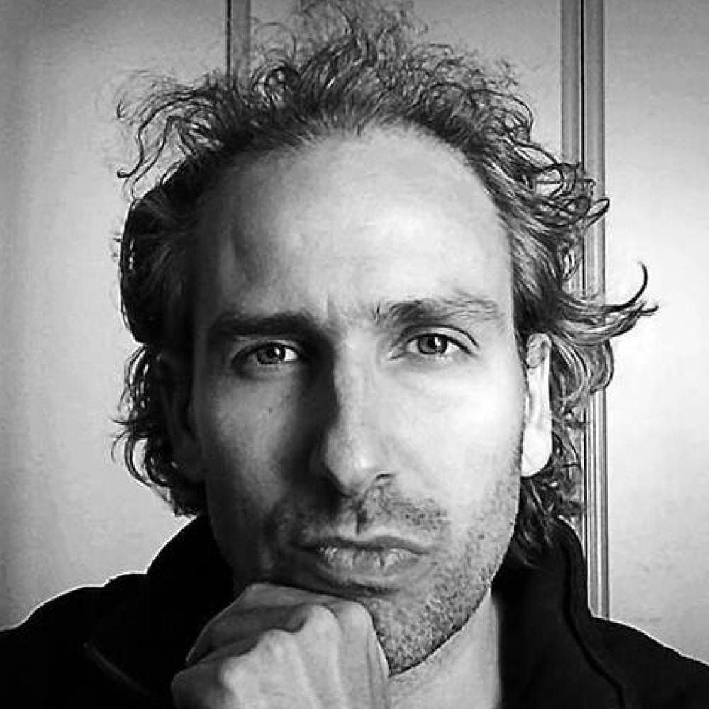 Portrait de Nicolas Beuglet, auteur français de thrillers, souriant devant une pile de ses romans.